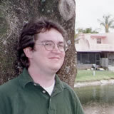 photo of Isaac Dealey, a young white  man with glasses and medium length hair resting against a tree trunk
