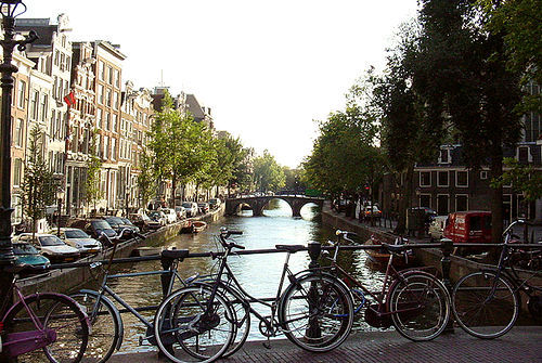 forground a bridge with two bicycles, a canal runs the the center of the picture, to the left a row of buildings, to the right a row of trees; the scene is from the Netherlands
