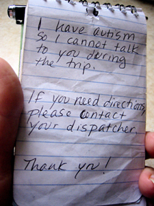 closeup of a hand holding a small notepad, on the notepad is written, \'I have autism so I cannot talk to you during the trip. If you need directions please contact your dispatcher.  Thank you!\'