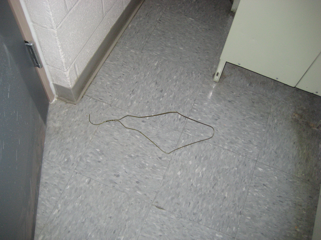 A used bent coat hanger on a dirty bathroom floor by a stall. Looks strikingly like a barrarcks bathroom.