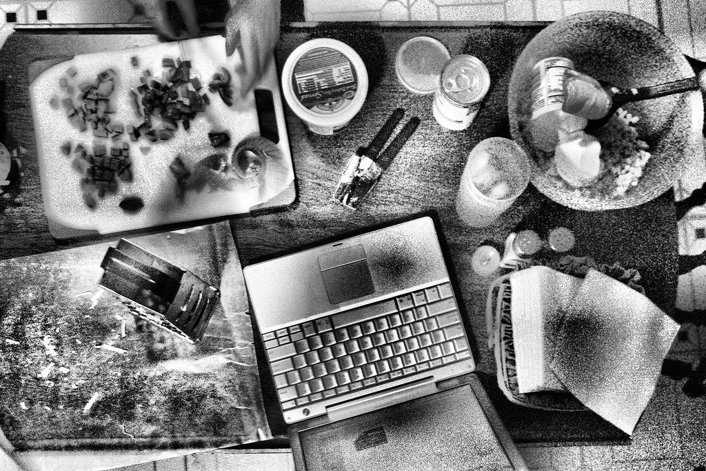 Black and white image of a kitchen countertop during meal preparation with food and garbage strewn about, and a laptop computer.