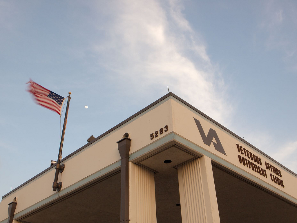 A lower shot of the corner of a tan building trimmed in brown. The roof holds an U.S. Flag waving in the wind, and the side says 
