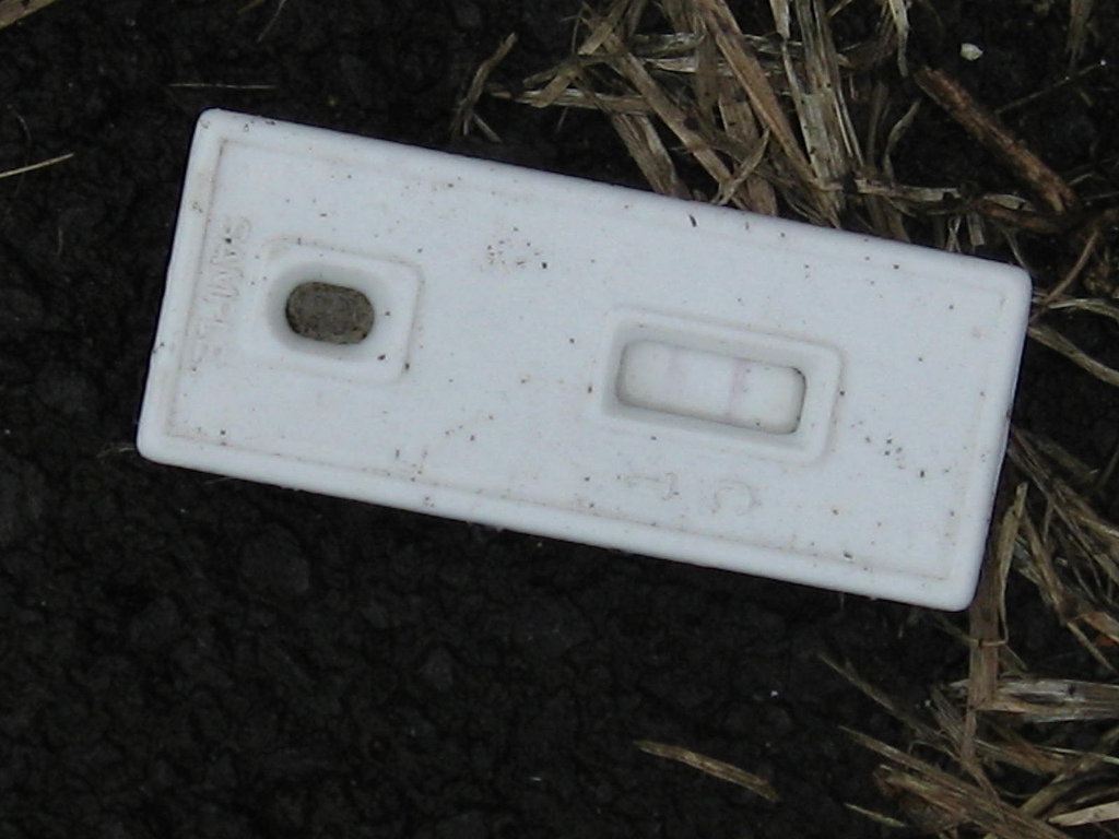 An old, white, battered positive pregnancy test lies forgotten on the ground.