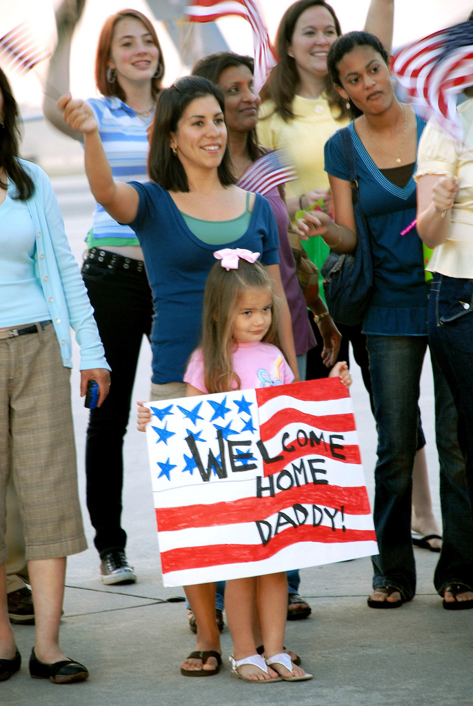 Several women and a small girl wave U.S. flags behind a posterboard painted to look like a U.S. flag. The board reads 