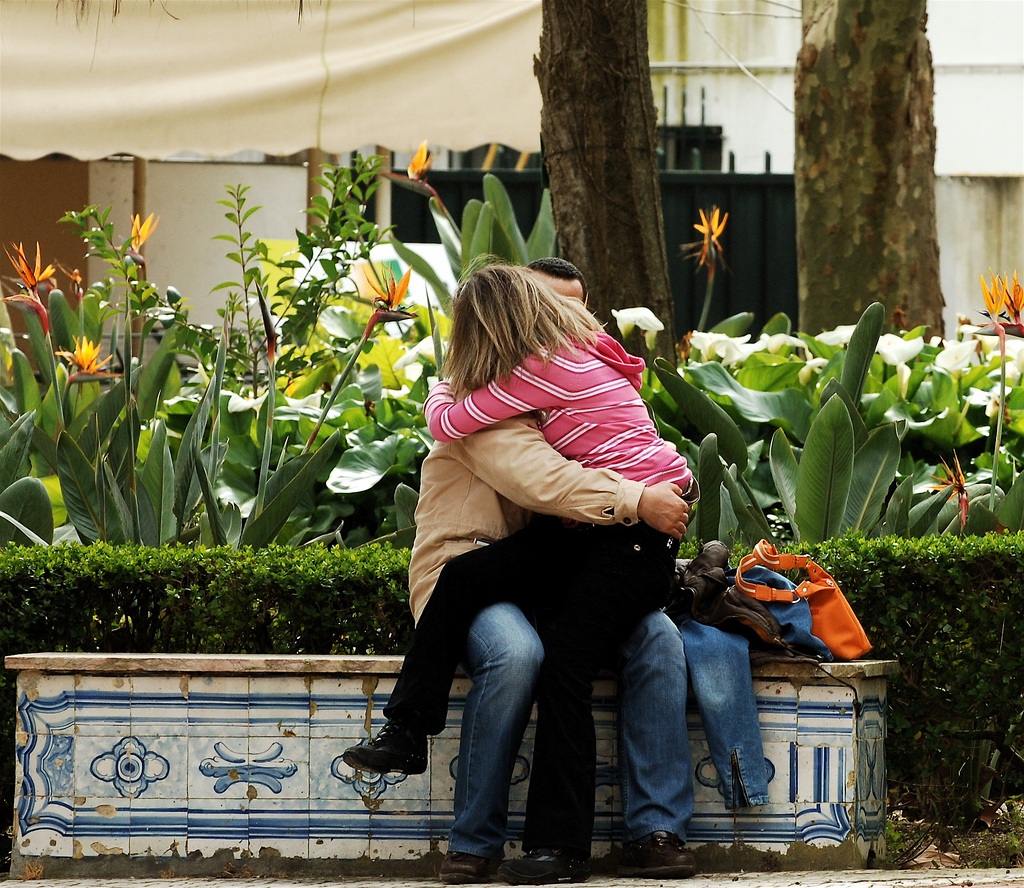 A seemingly white man and indistinguishable but blonde woman sit on a blue and whilte tiled flower box filled with birds of paradise, engaged in a deep kiss.