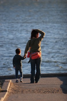 A pale woman in a sweater and cargo pants holds the hand of a pale boy in short pants and a sweatshirt. She absently runs her hand through her wind-tossed hair as they wander down a pier towards a body of water.