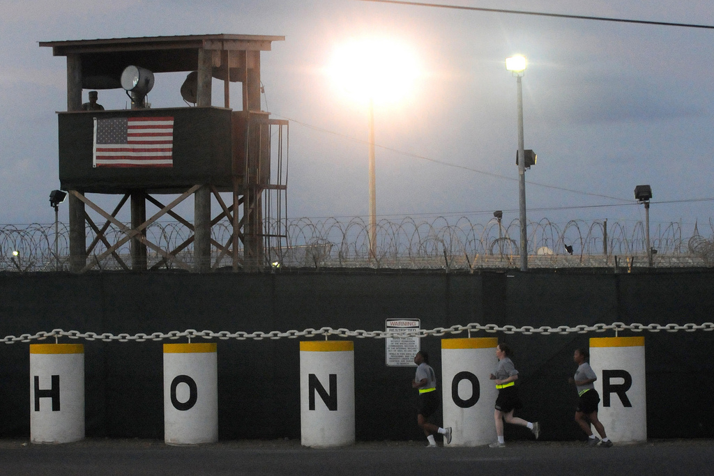 A military guard shack with extremely bright flood lights and wide loops of concertina wire over barbed wire sits atop a wall of colums, each with a liter, H-O-N-O-R. Three women in Army PT gear (grey t-shirts and black shorts with reflective belts) jog past.