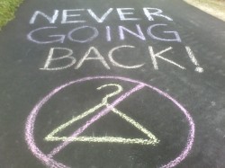 A chalk drawing on pavement, of a hanger circled and crossed out. Text reads "NEVER GOING BACK!".